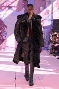 Male model walking a fashion runway in a dark, fur-trimmed coat and high boots, posing confidently amid pink murals.