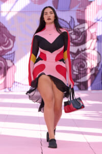 Female model walking a runway in a color-block pink, black and red dress with sheer sleeves, carrying a small red and black handbag.