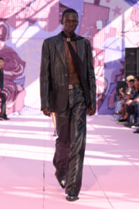 Male model on a pink runway wearing a black leather blazer and pants, no shirt underneath, with a black scarf and jeweled brooch at the neck.