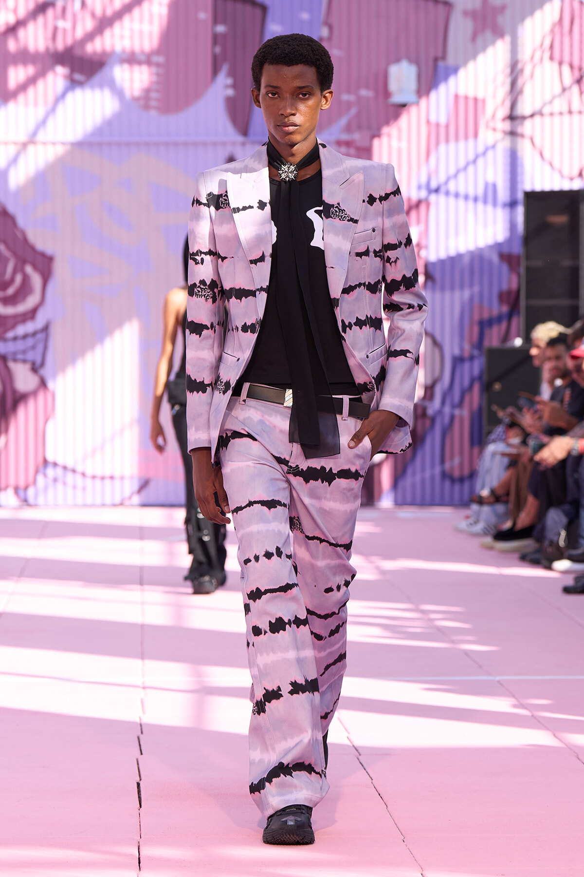 Male model walks a fashion runway in a pink and black tie-dye suit with a black necktie and silver brooch accessory.