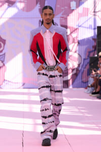 Male model walks a pink-themed runway in a color-blocked shirt (lavender sleeves, red panels) with a pink tie and tie-dye pants, accessorized by a jeweled belt.