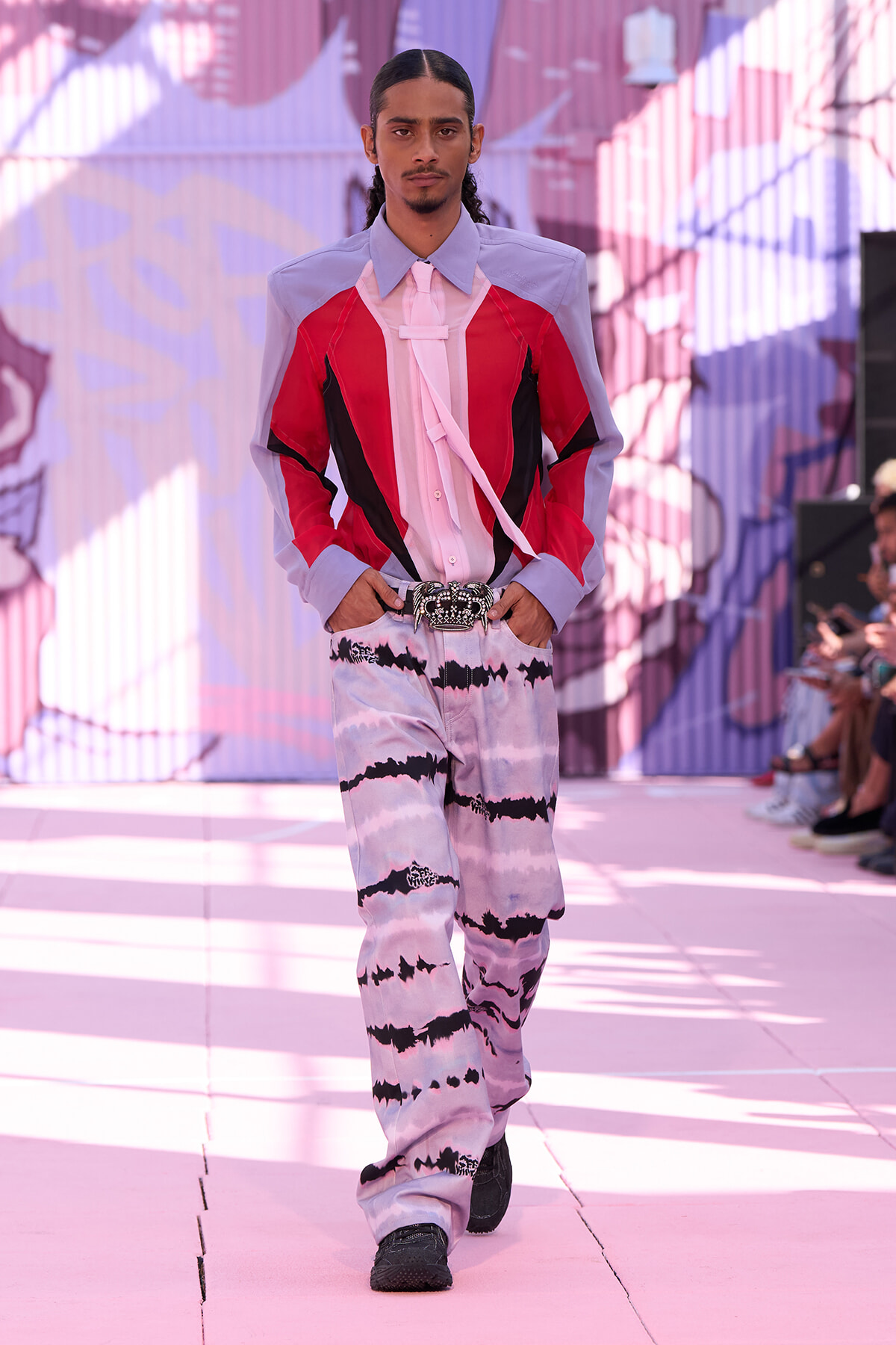 Male model walks a pink-themed runway in a color-blocked shirt (lavender sleeves, red panels) with a pink tie and tie-dye pants, accessorized by a jeweled belt.