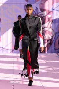 Female model on a pink outdoor runway wearing a black tailored long coat with color-block sleeves and a fringed hem, revealing bright patterned pants.