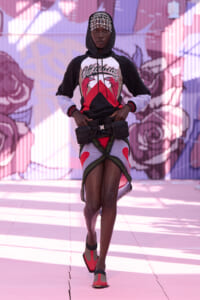 A fashion model walks toward the camera in a black-and-white hoodie, decorative headpiece, and a utility belt over a colorfully wrapped mini skirt, with a mural backdrop.