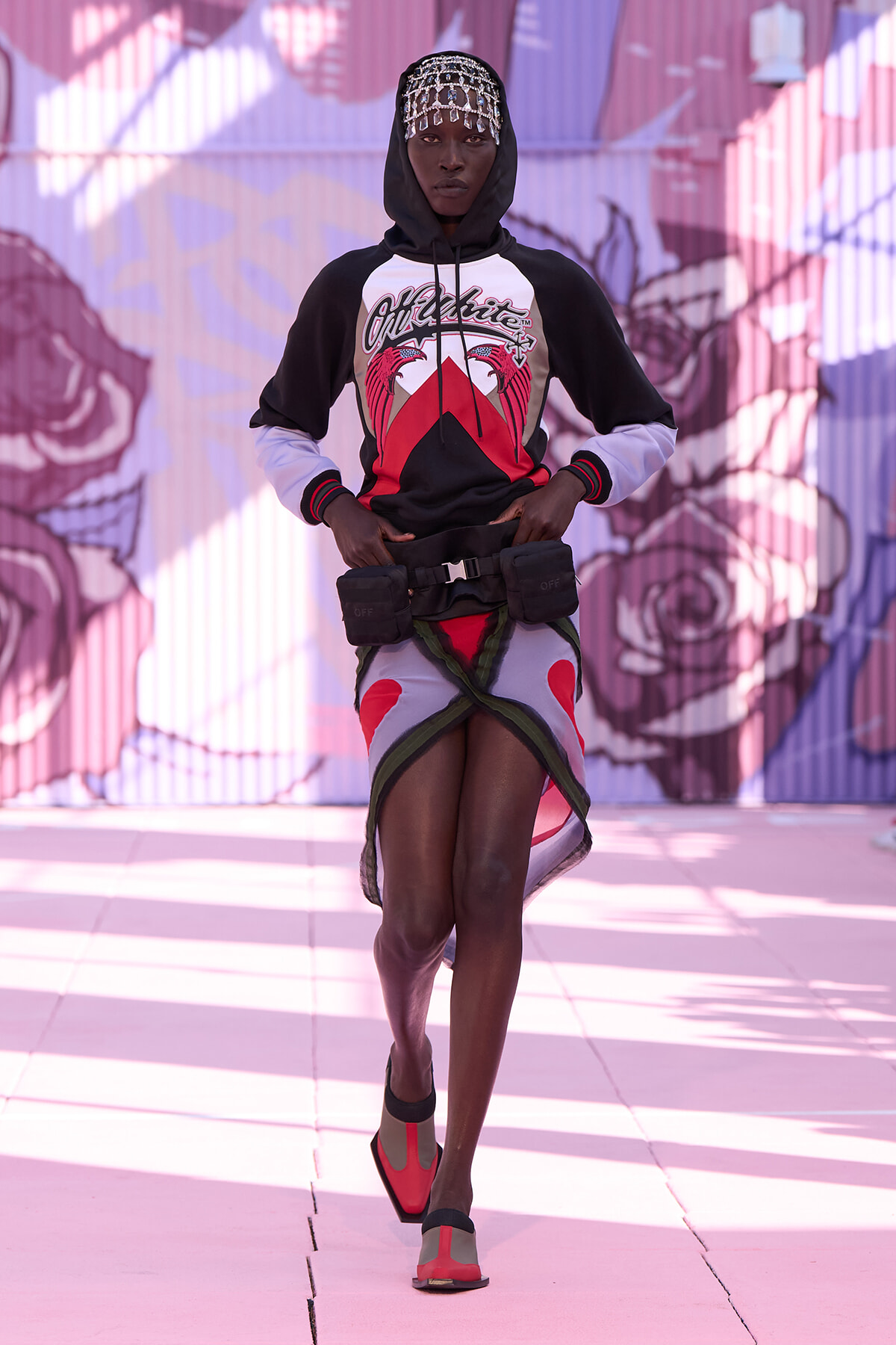 A fashion model walks toward the camera in a black-and-white hoodie, decorative headpiece, and a utility belt over a colorfully wrapped mini skirt, with a mural backdrop.