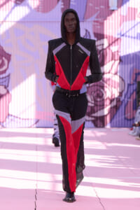 Model walking the runway in a black, red, and grey color-blocked hooded jumpsuit with geometric panels against a mural backdrop.