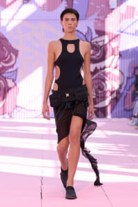 Model walking a fashion show in a black cutout bodysuit with a cargo belt of pouches, against a pink graffiti backdrop.