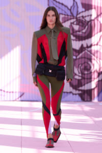 Model walking a fashion runway in a color-blocked olive, red, and black outfit with a belt bag at the waist, graffiti background behind.