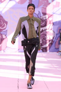 Model walks a fashion runway in a utilitarian olive shirt with color-block panels, a beige tie, and a black-and-beige cargo-panted outfit with a belt bag.