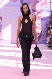 Runway model wearing a black hooded halter top with a circular cutout and cargo pants with utility pockets, carrying a black bag on a pink-lit runway.