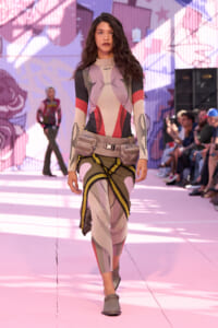 Model walks the pink runway in a geometric color-block bodysuit with a beige utility belt and taupe shoes.