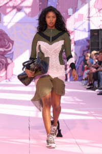 Model walks a fashion show wearing a color-block crochet top with green, pink, and black panels, olive skirt, and oversized bag accessory.