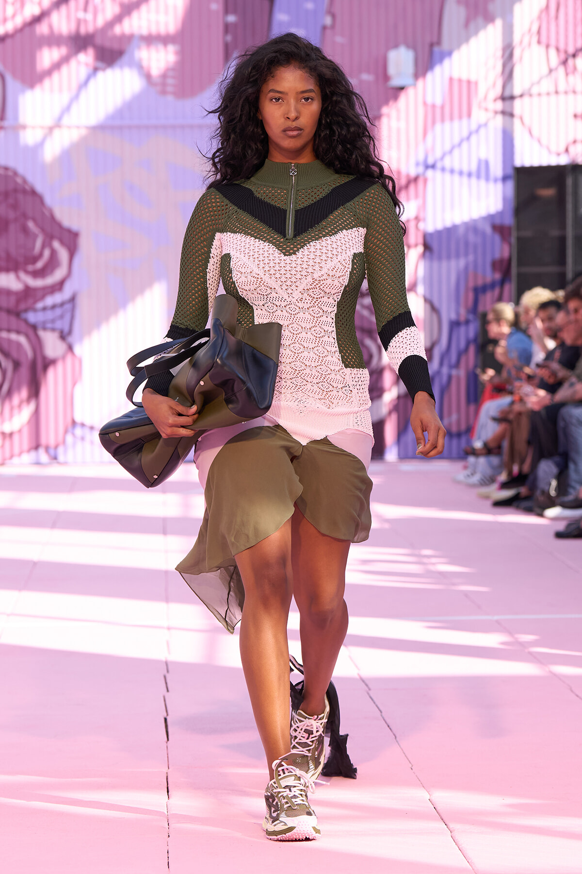 Model walks a fashion show wearing a color-block crochet top with green, pink, and black panels, olive skirt, and oversized bag accessory.