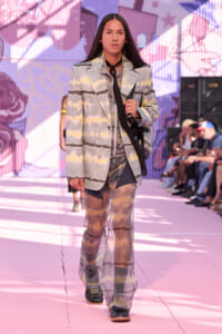 Model walking a runway in a pastel tie-dye suit with a black neck scarf, carried bag strap over shoulder, pink stage backdrop behind.