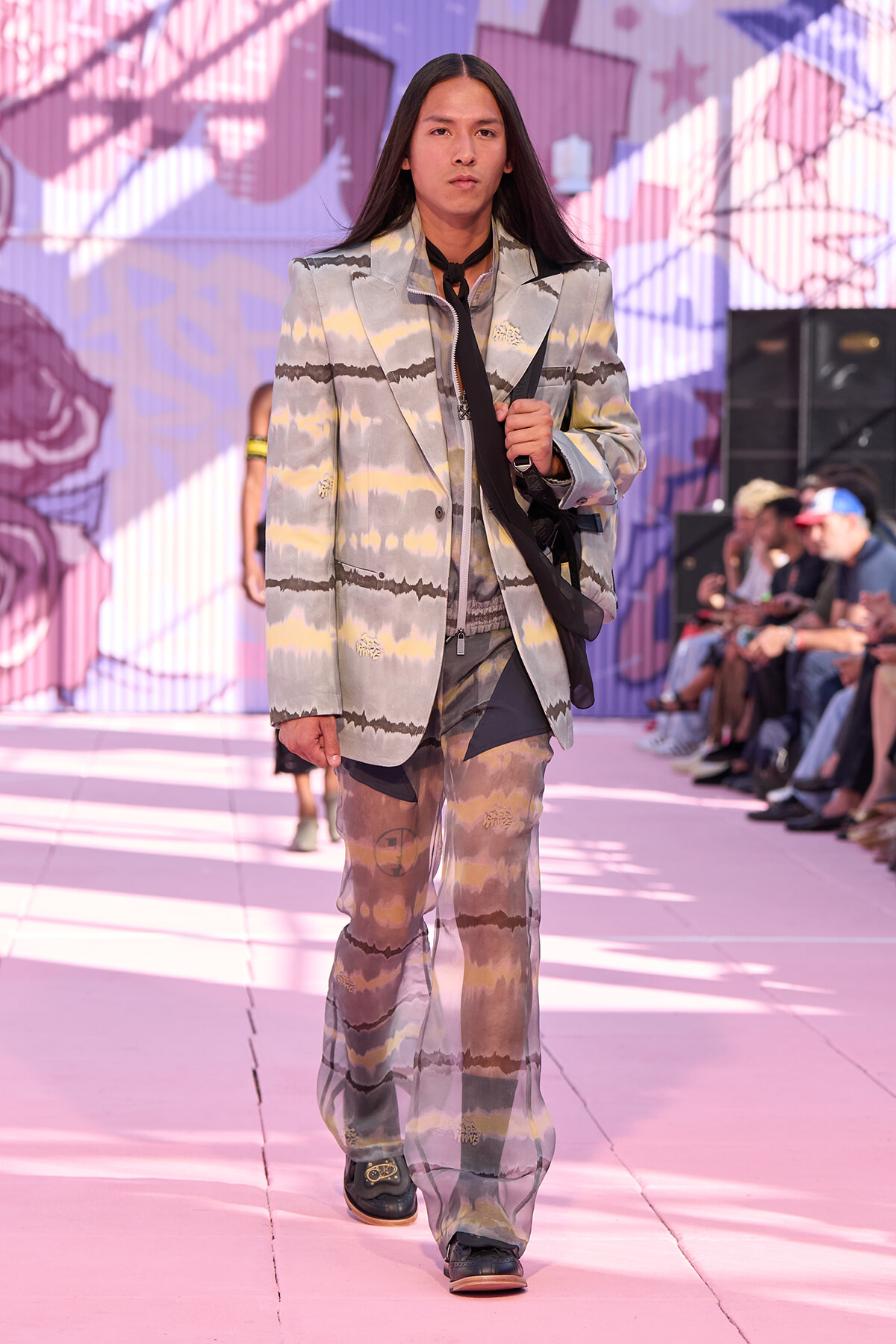 Model walking a runway in a pastel tie-dye suit with a black neck scarf, carried bag strap over shoulder, pink stage backdrop behind.