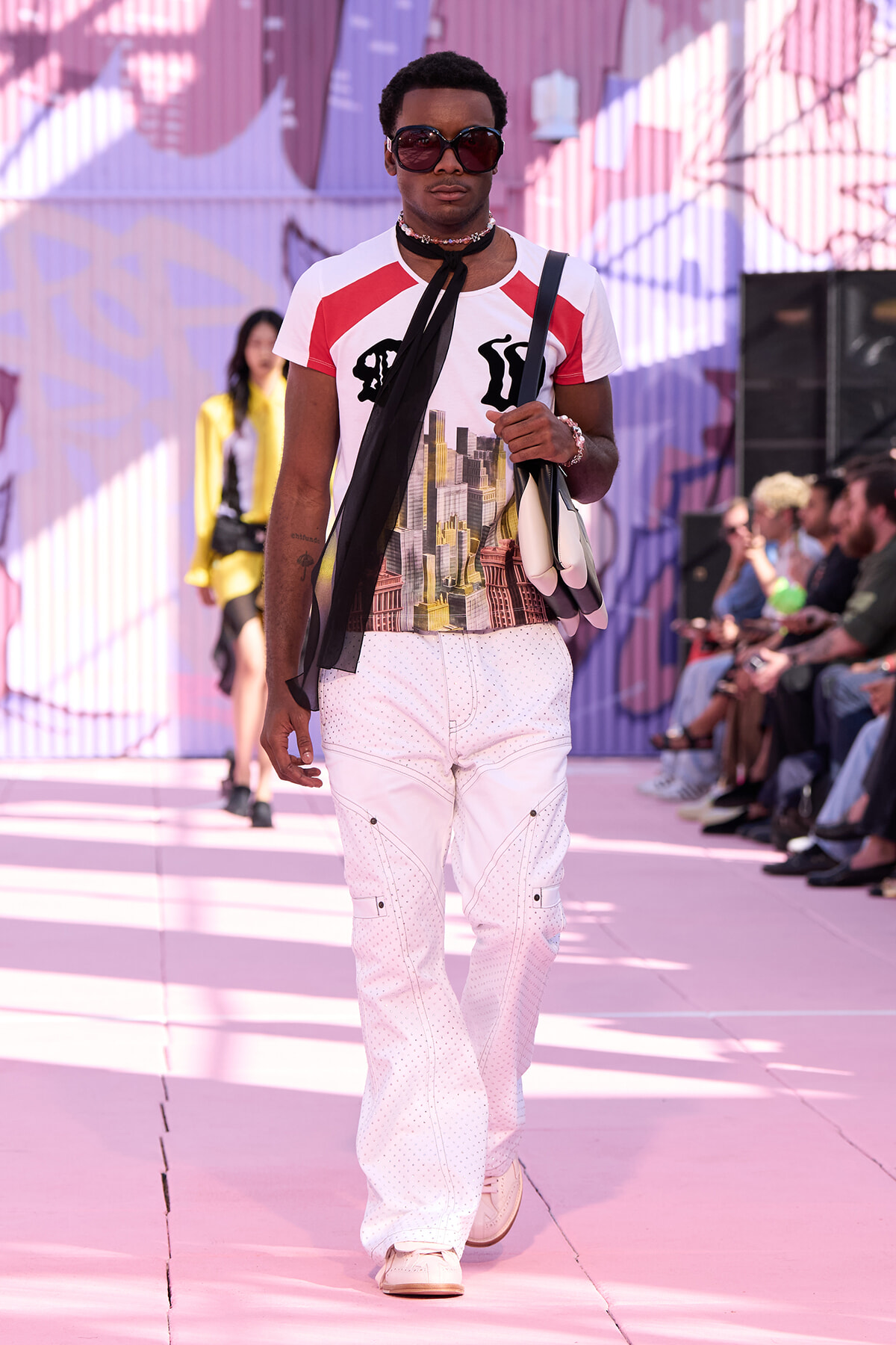 Male model walks a pink-lit runway in a graphic cityscape tee, white dotted cargo pants, large sunglasses, and a scarf draped around the neck, carrying a bag over the shoulder amidst cheering onlookers.
