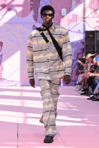 Male model walking a pink-toned runway in a pastel tie-dye suit and dark sunglasses, with a black crossbody strap across his chest.