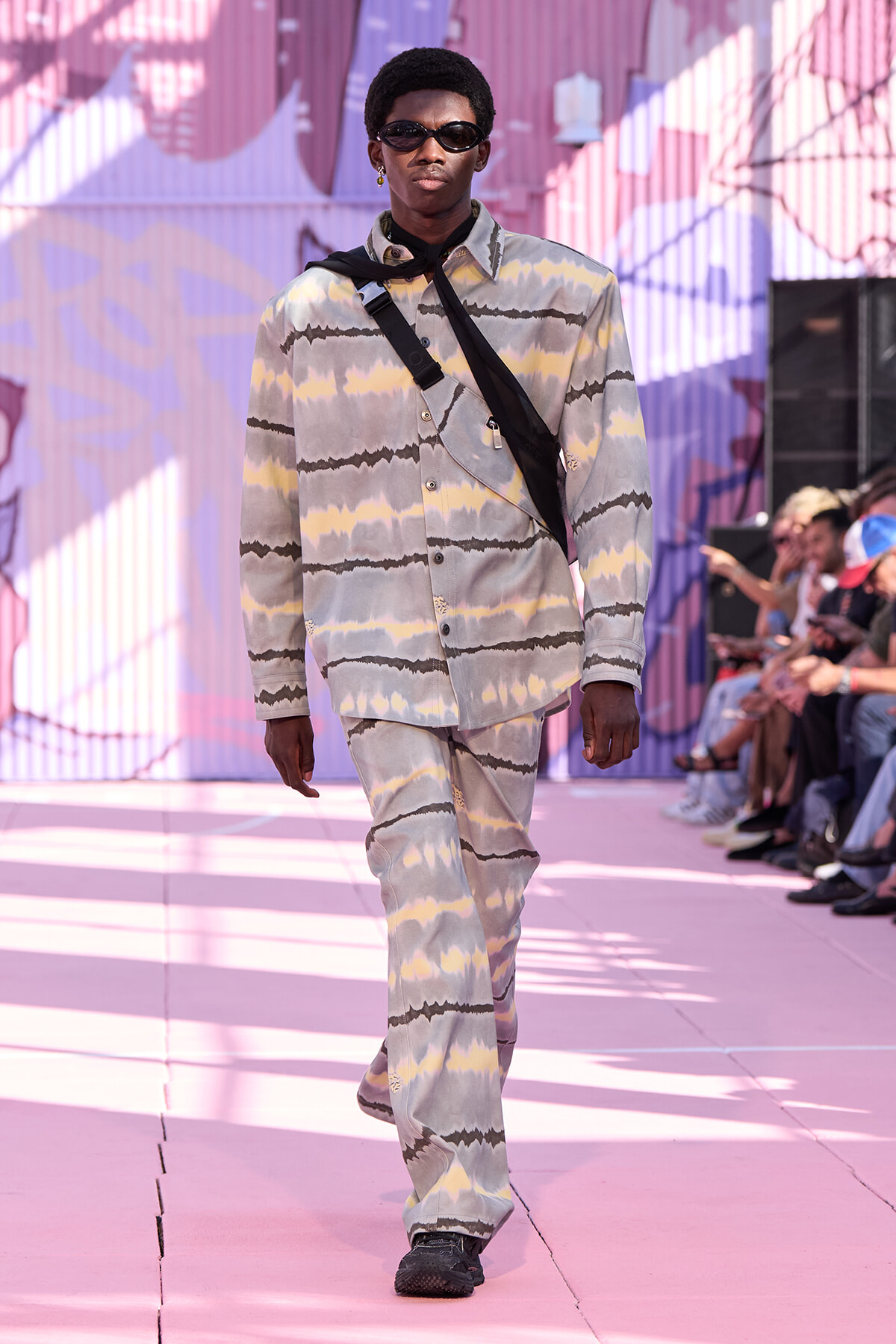 Male model walking a pink-toned runway in a pastel tie-dye suit and dark sunglasses, with a black crossbody strap across his chest.