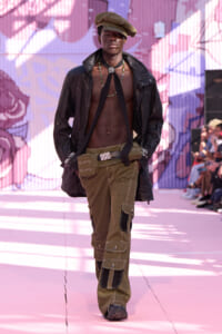 Male runway model walking the catwalk in an open black leather jacket, olive cargo pants, bare chest with bead necklaces and a brown cap, pink mural backdrop behind him.
