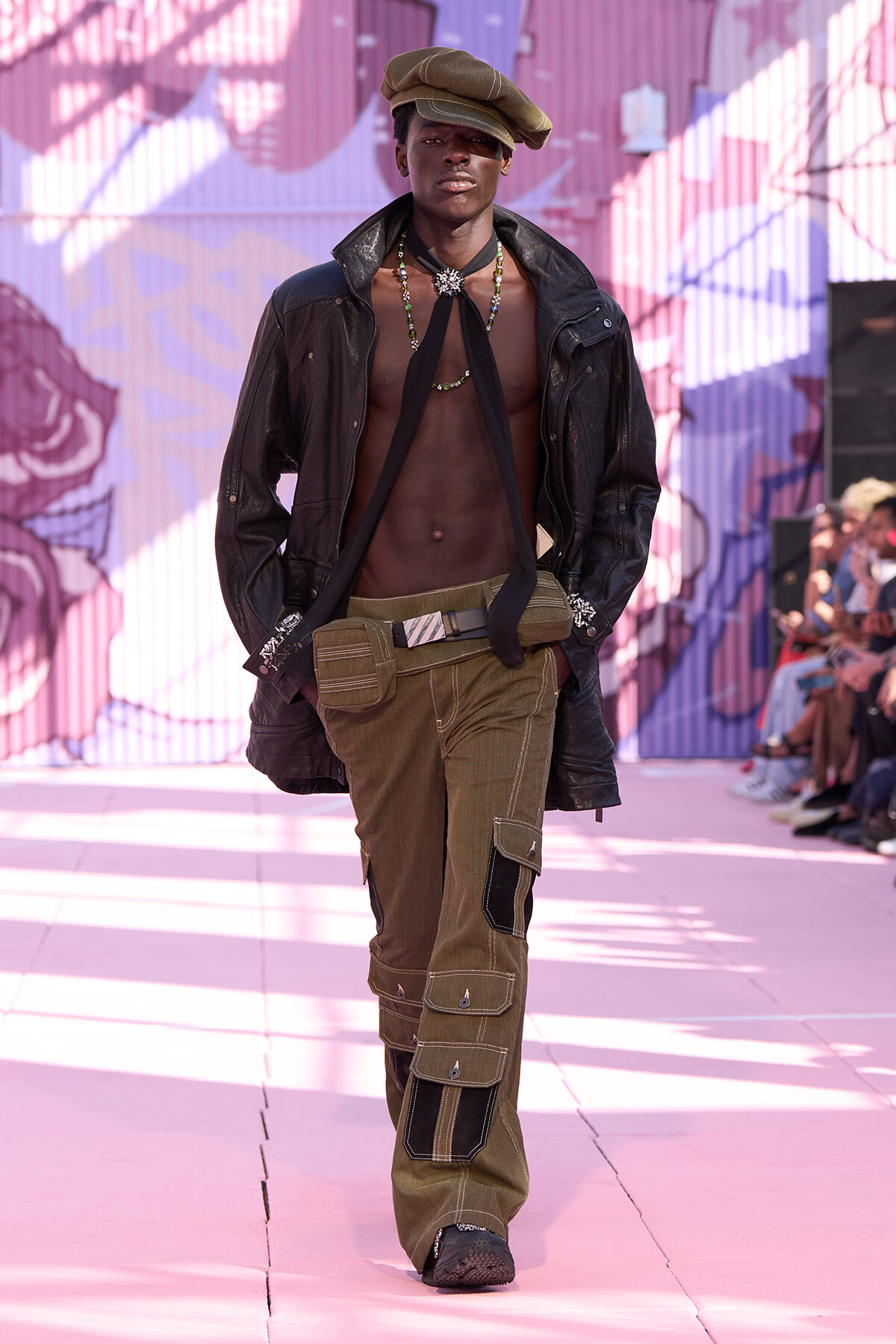 Male runway model walking the catwalk in an open black leather jacket, olive cargo pants, bare chest with bead necklaces and a brown cap, pink mural backdrop behind him.