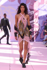 Model walking the runway in a pink patterned halter dress with a high-low hem and dark arm wraps.
