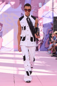 Male model walking a pink-lit runway in a white and black sleeveless utility jumpsuit, oversized sunglasses, holding a black scarf and accessories.