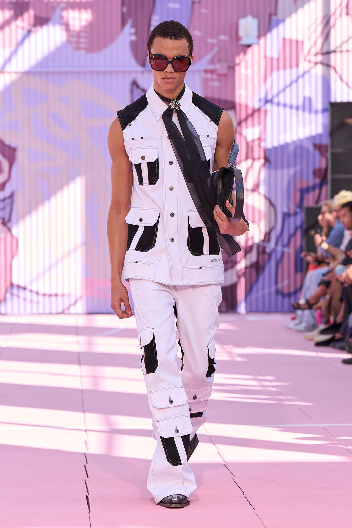Male model walking a pink-lit runway in a white and black sleeveless utility jumpsuit, oversized sunglasses, holding a black scarf and accessories.