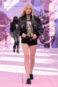 Model walking a runway in a black embroidered bomber jacket, graphic top, and black shorts with utility pockets against a pink graffiti backdrop.