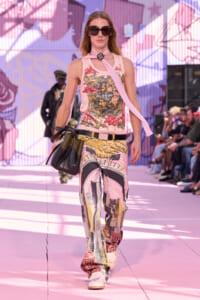 Model walking a pink runway in a graphic print sleeveless top, matching cityscape-print pants, a pink scarf, oversized sunglasses, and a black handbag.