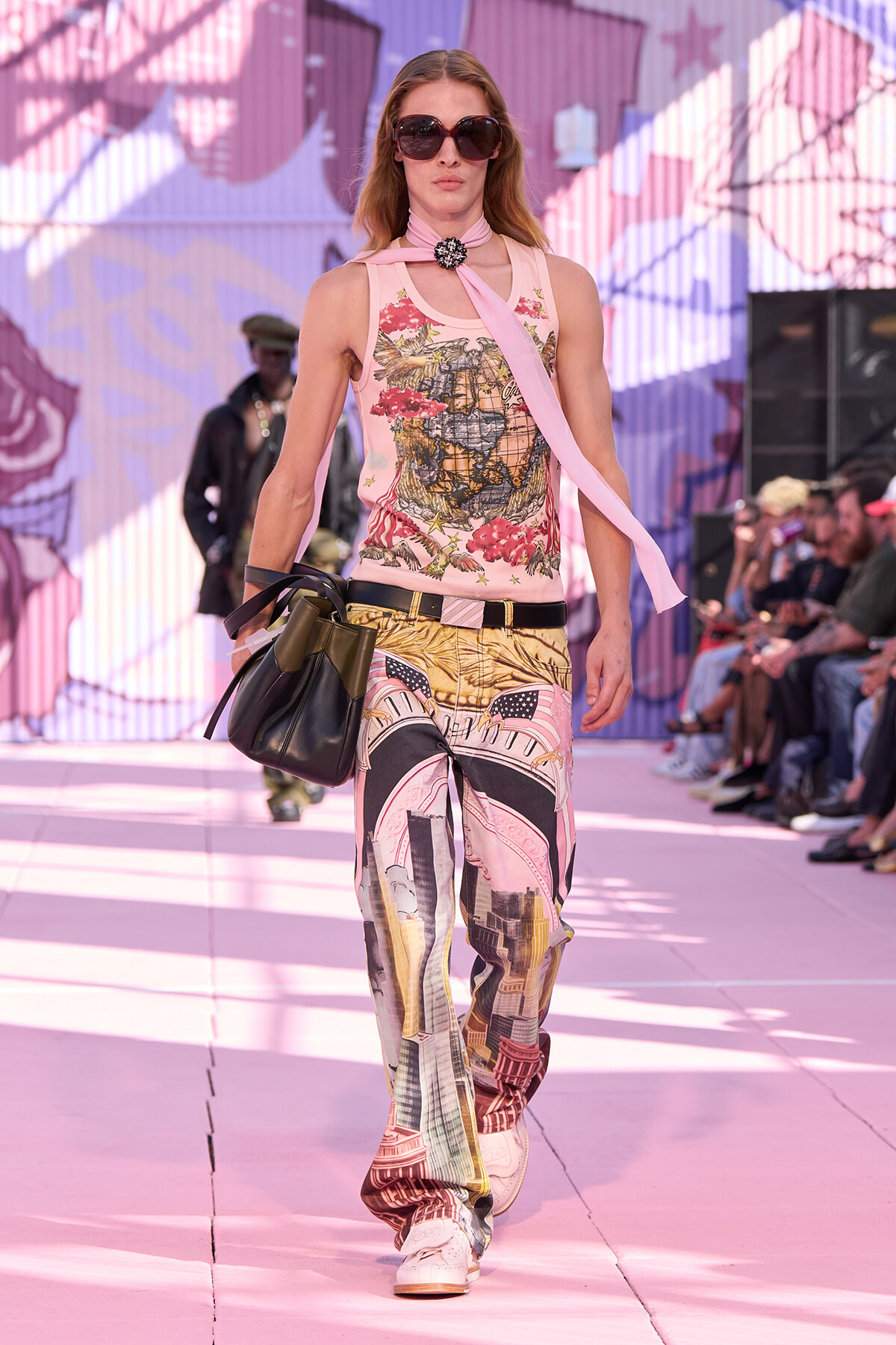 Model walking a pink runway in a graphic print sleeveless top, matching cityscape-print pants, a pink scarf, oversized sunglasses, and a black handbag.