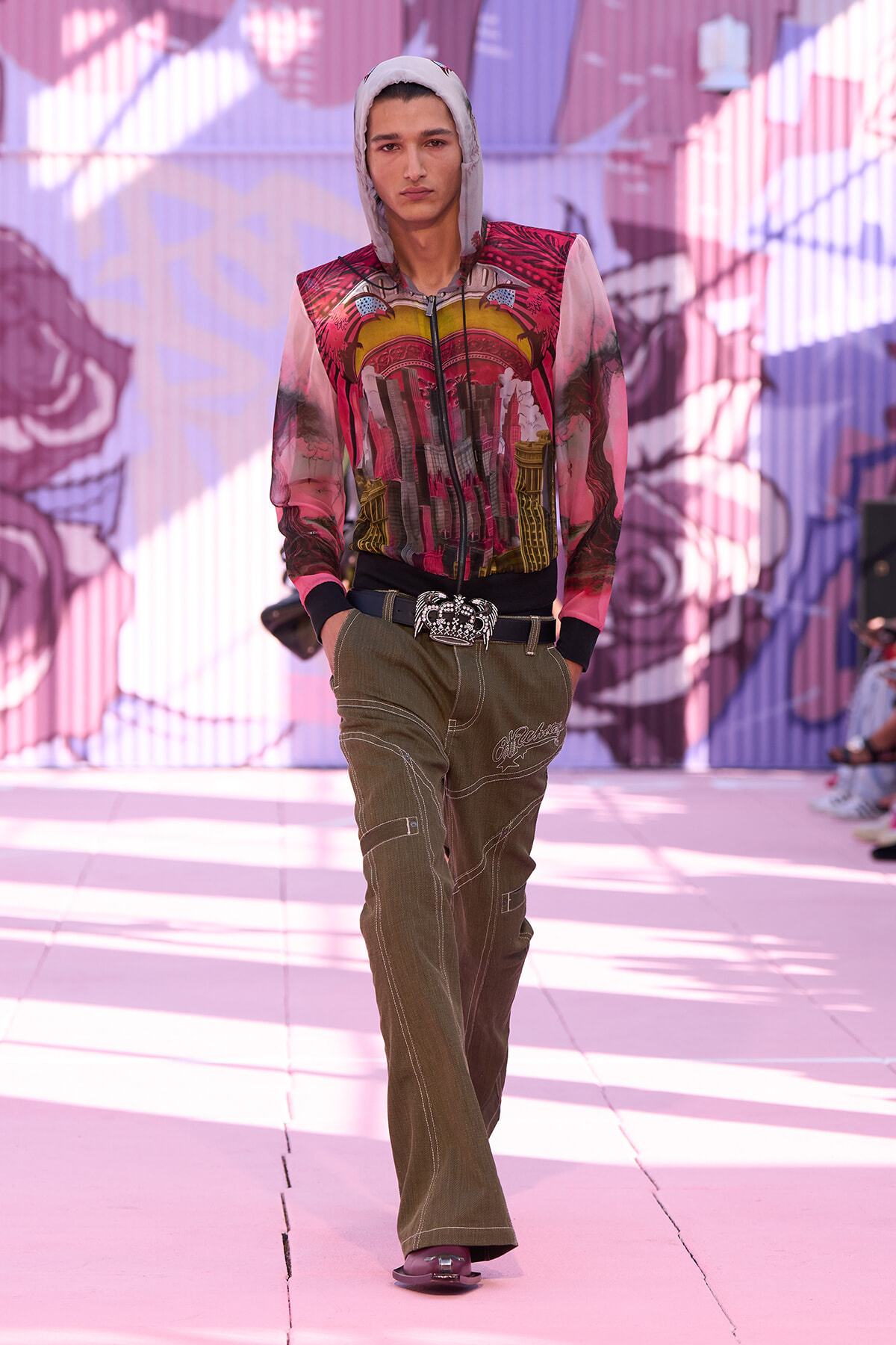 Male model walks the runway in a pink-red graphic hoodie, olive cargo pants, and a jeweled belt, with a graffiti backdrop.