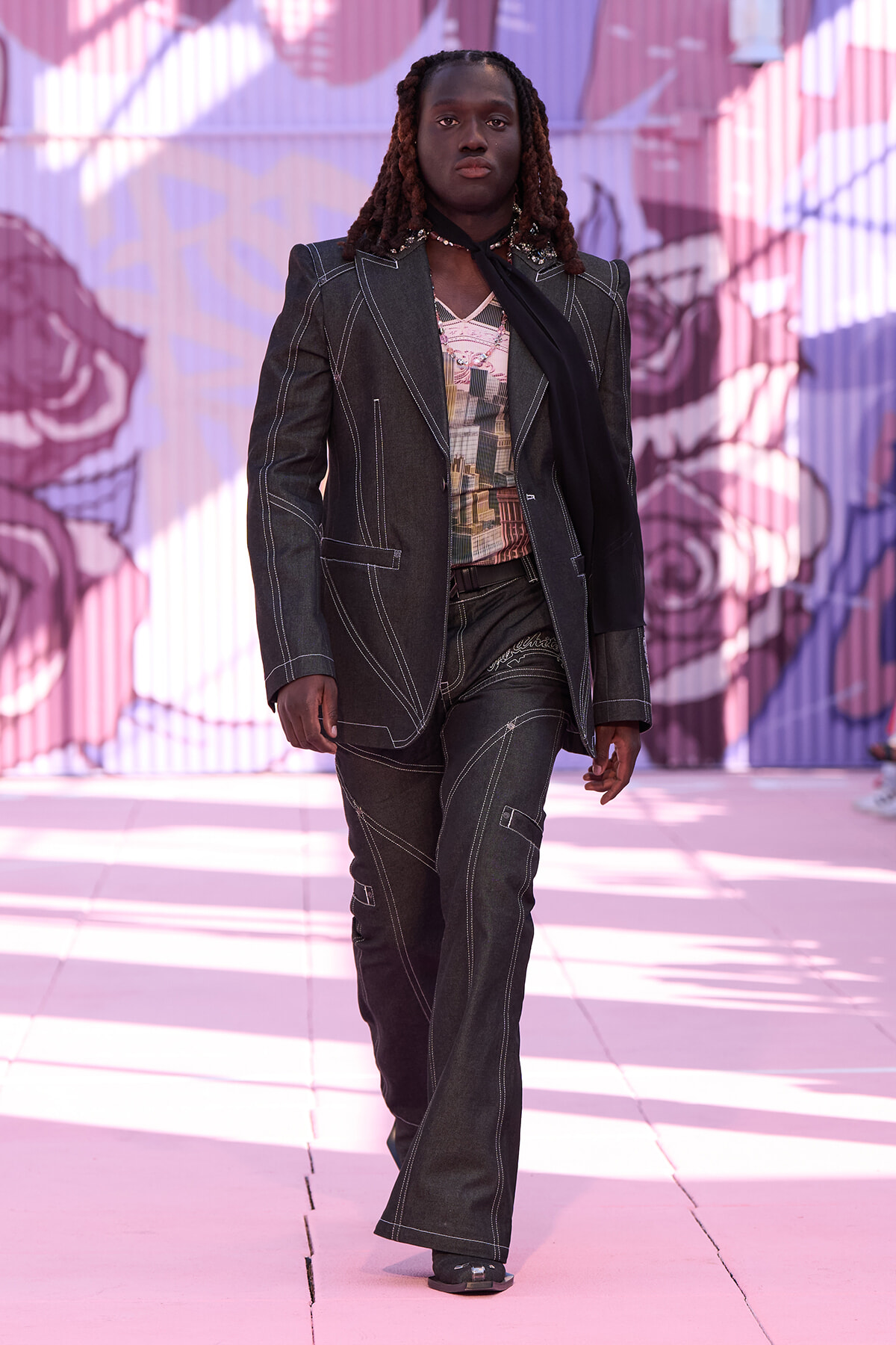 Runway model wearing a dark denim suit with white contrast stitching, a patterned top, and a black scarf, walking in front of a pink mural backdrop.