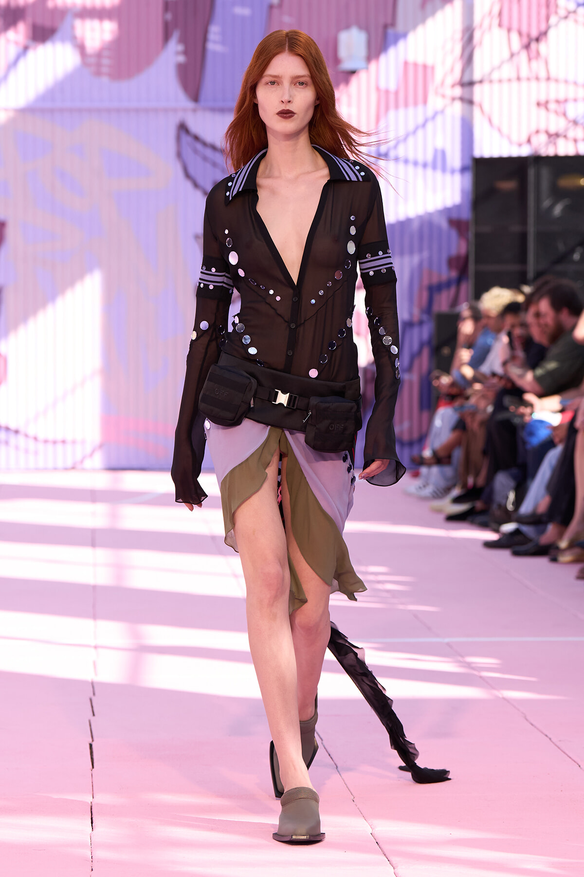 Model walking a pink runway in a sheer black blouse with metallic discs and a color-block wrap skirt, utility belt and pouches nearby.