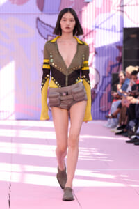 Runway model in olive-green top with yellow accents and studded detailing, paired with brown shorts and a gray utility belt bag.