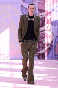 Male model walking a runway in an olive pinstripe suit with a jeweled collar and black top, against a colorful mural.