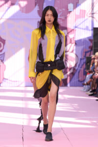 Model walking a fashion runway in a yellow, gray, and black color-blocked blouse and matching asymmetric yellow skirt with a black belt and utility pouches. Colorful mural backdrop.