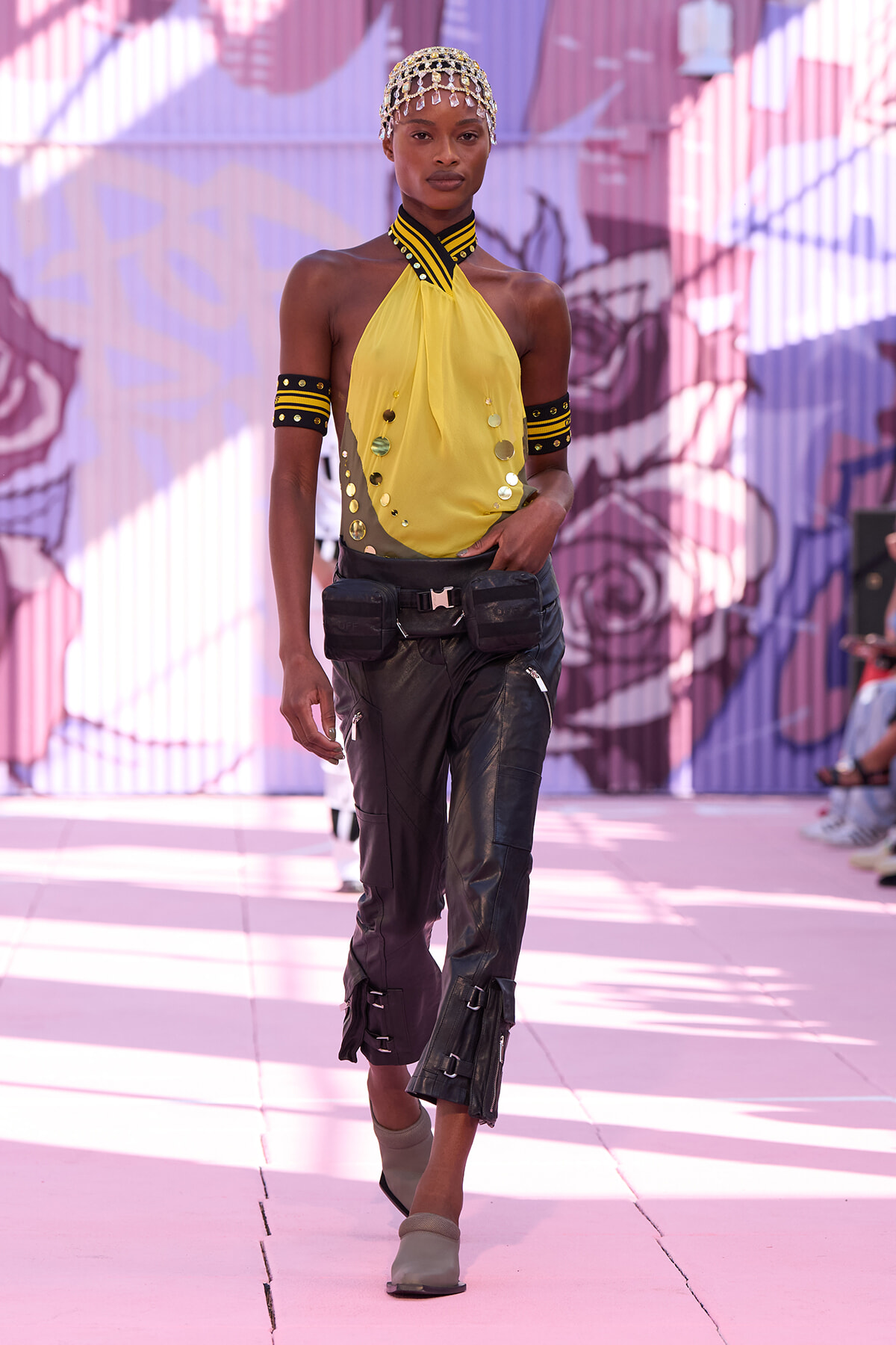 Model on a runway wearing a yellow halter top, black cargo pants with buckles, beaded headpiece, and black-and-yellow armbands.