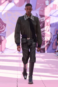 Male model walking a runway in an olive jacket with a jeweled bold collar, black scarf, dark jeans with prominent white stitching, and tall black boots against a pink graffiti backdrop.