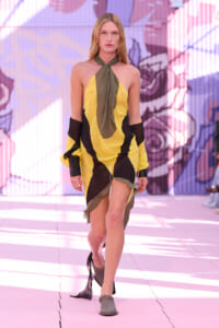 Model walking a fashion-show runway in a yellow and black color-block dress with an olive-green scarf tied at the neck.