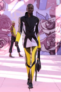Model walking a fashion show in a color-block blazer with white panels and bright yellow leggings, carrying a black handbag on a pink mural backdrop.
