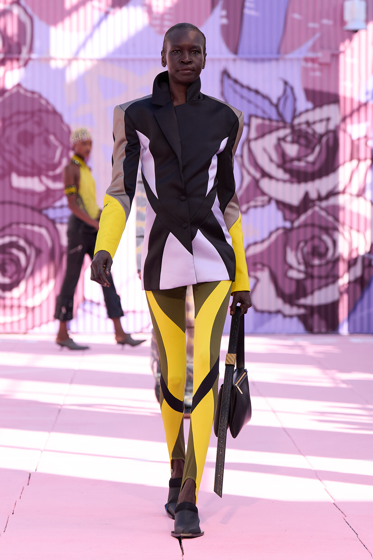 Model walking a fashion show in a color-block blazer with white panels and bright yellow leggings, carrying a black handbag on a pink mural backdrop.