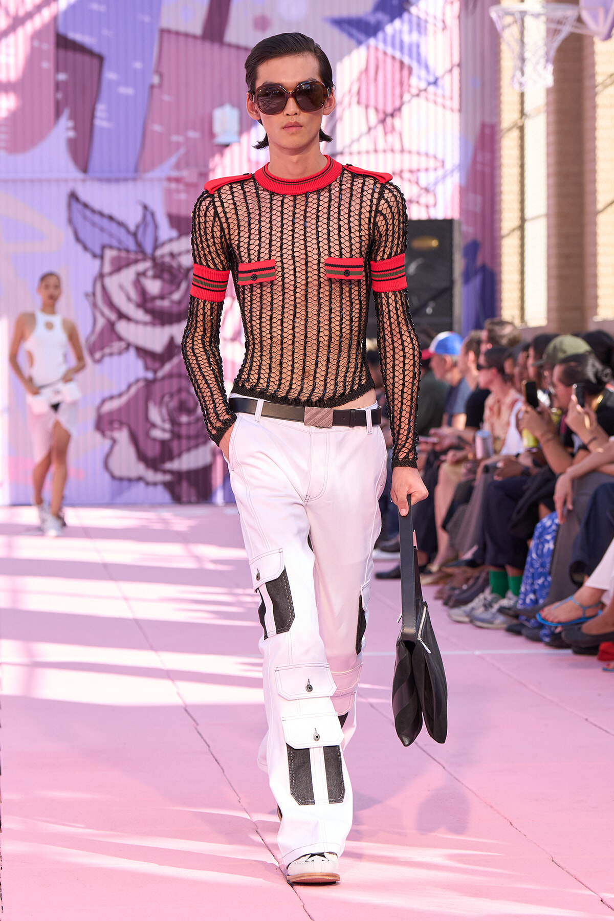 Model walking a pink runway in a black mesh top with red trim and white cargo pants, wearing sunglasses and carrying a black bag.