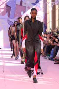 Model walks a pink runway in a black satin coat with red and white color-block sleeves and fringe at the hem; audience watches from sides.