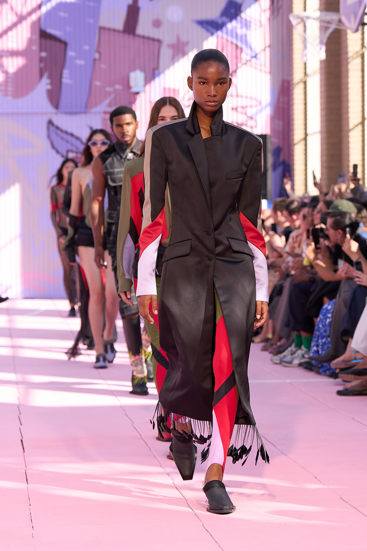 Model walks a pink runway in a black satin coat with red and white color-block sleeves and fringe at the hem; audience watches from sides.