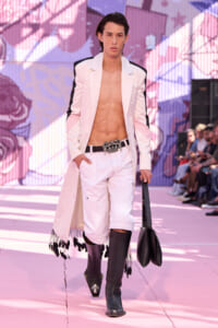 Male model walking runway in a pale pink and black suit with no shirt, white shorts, and tall black boots on a pink graffiti backdrop, carrying a black bag.