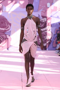 Model walking a runway in a light pink halter dress adorned with reflective circles and black-and-white striped accents, against a colorful mural backdrop.