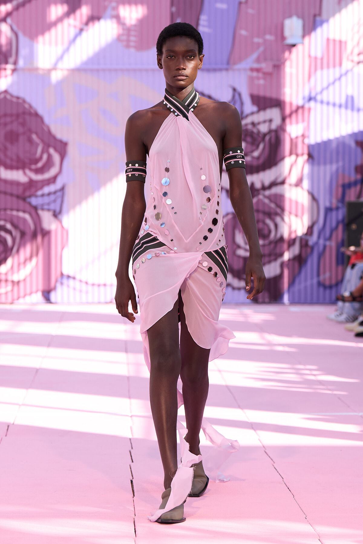 Model walking a runway in a light pink halter dress adorned with reflective circles and black-and-white striped accents, against a colorful mural backdrop.