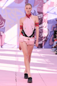 Model walking a fashion runway in a pink halter top with silver sequins, matching pink shorts and belt bags.