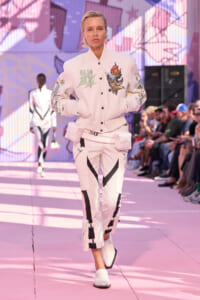 Model walking the runway in a white varsity jacket with embroidered eagle and torch patches, pink-toned set design in the background.