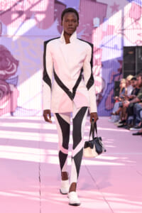 Model on a pink runway wearing a geometric pink and black blazer and leggings, carrying a black-and-cream handbag.